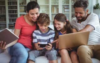 Photo of a family sitting on a couch using a laptop, phone, and tablet